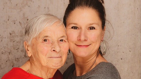 Eine Mittvierzigerin hält ihre Großmutter fest im Arm. Beide Frauen blicken lächelnd in die Kamera.