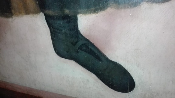 Auf einem Gemälde sieht man von ganz nahem fotografiert einen linken Fuß in einem dunklen Lederschuh stecken. Das Detail ist eine Aufnahme eines Cranach-Bildes aus dem Dom zu Meißen.