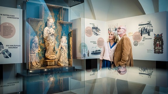 Eine Frau mit weißem Haar und ein Mann mit Glatze blicken sich in einem Ausstellungsraum mit Glasvitrinen und Erklärtafeln um.