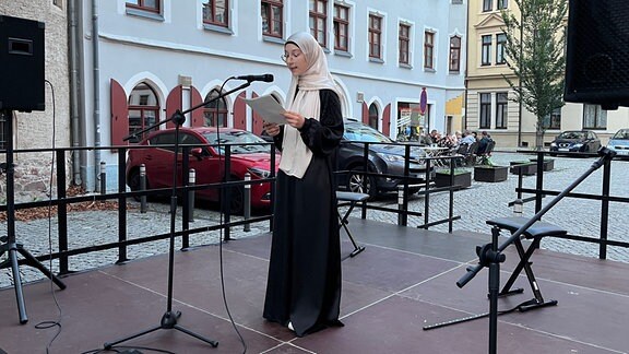 Eine junge Frau mit einem Kopftuch steht vor einem Mikrofon und spricht von einer Bühne.