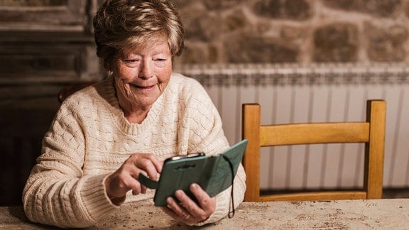 Eine ältere Frau sitzt mit einem Smartphone am Tisch.