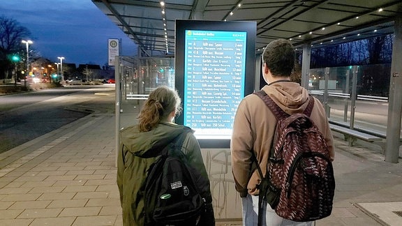 Pendler stehen am Busbahnhof in Meißen vor einer Informationsanzeige.