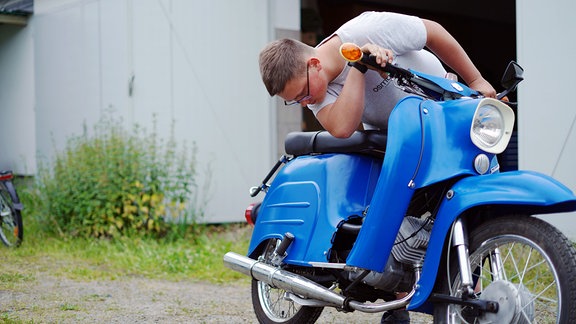 Ein Junge mit Brille sitzt auf einem blauen Moped.