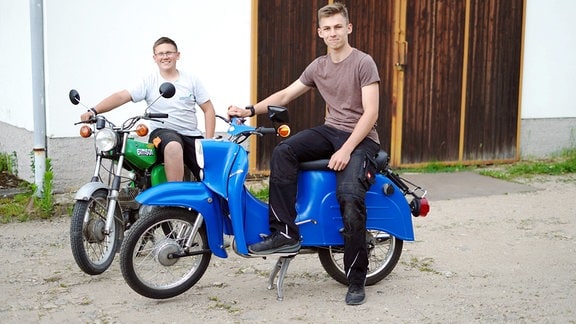 Ein Junge sitzt auf einem grünen und ein anderer auf einem blauen Moped.