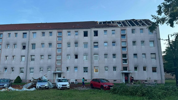 Ein DDR-Neubaublock ist am Dach stark beschädigt. Balken und Dachpappe liegt auf Autos, die schwer beschädigt sind.