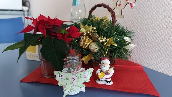 Auf einem Tisch stehen auf einer roten Serviette ein kleiner Weihnachtsstern im blumentopf, ein Gesteck und ein eine Mini-Figur in Form eines Weihnachtsmannes.