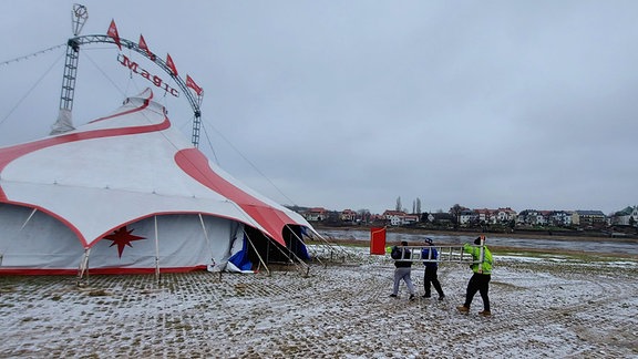 Auf einer Wiese steht ein Zirkuszelt. An einem grauen Wintertag laufen drei Männer in Arbeitssachen am Zelt entlang. Zwei tragen eine große Leiter auf ihren linken Schultern.