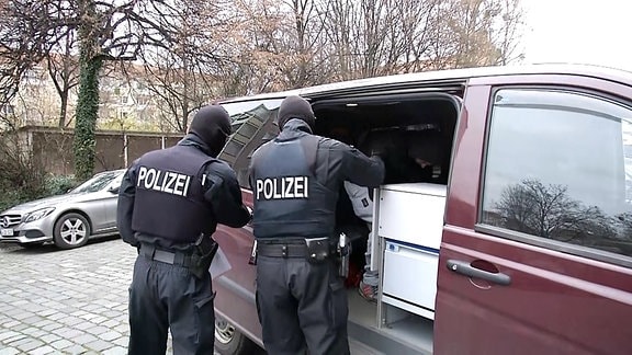 Ein Gefangener sitzt in einem Kleinbus der Polizei. Zwei maskierte Polizisten stehen an der geöffneten Seitentür.