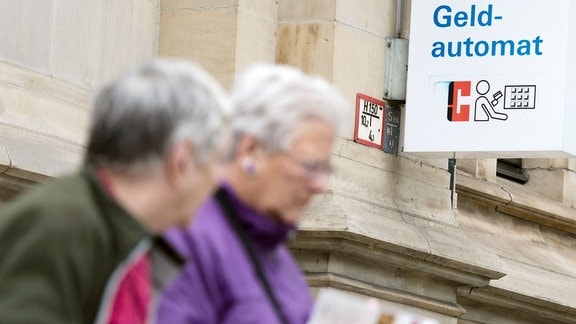 Zwei Seniorinnen gehen an einer Bank mit einem Schild "Geldautomat" vorbei. 