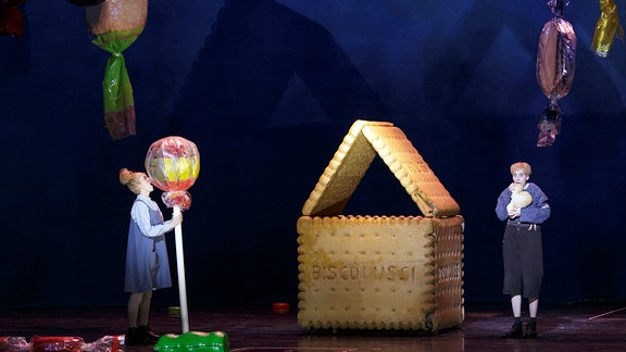 Hänsel und Gretel, ein Haus aus riesengroßen Keksen, von oben hängen große Bonbons, ein Mädchen leckt an einem riesigen Lolli, ein Junge steht daneben.