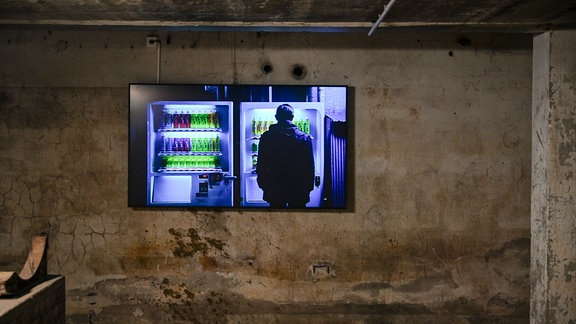 Auf einem Fernseher in einer dunklen Umgebung ist eine Filmszene zu sehen, die eine Person vor leuchtenden Getränkeautomaten zeigt. Den Film halt Carsten Nicolai gedreht. 