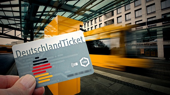 Eine Hand hält ein Deutschlandticket, im Hintergrund fährt eine Straßenbahn an einer Haltestelle