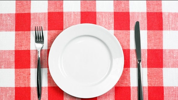 Ein leerer weißer Teller und Besteck auf einem rot-weiß karrierten Tischtuch.
