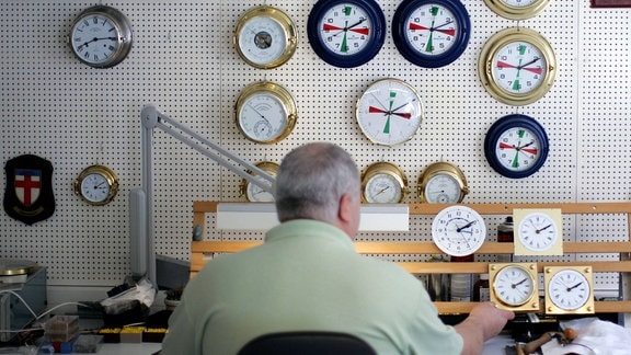Ein Uhrmacher des Unternehmens Mühle Glashütte an seinem Arbeitsplatz, vor ihm an der Wand hängen Uhren in unterschiedlichen Größen.