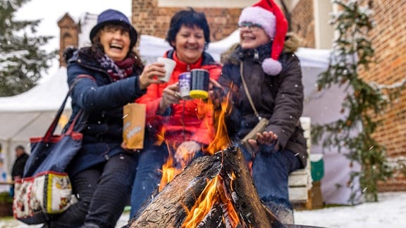 Drei illustre Damen stoßen auf dem Weihnachtsmarkt in Leuthen, an einem Feuer sitzend, mit Glühwein an.