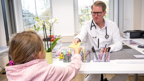 Ein Hausarzt übergibt in einem Sprechzimmer seiner Praxis einem jungen Mädchen den Impfausweis.