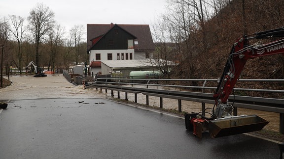 Ein Bagger steht auf einer überfluteten Straße neben einem Wohnhaus, das von Wasser umgeben ist.