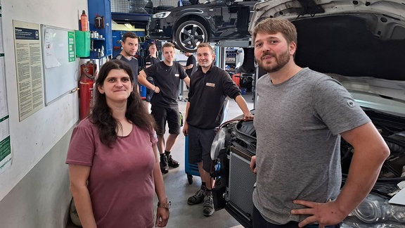 Eine Frau und weitere Mitarbeiter der Autowerkstatt lächeln in die Kamera.