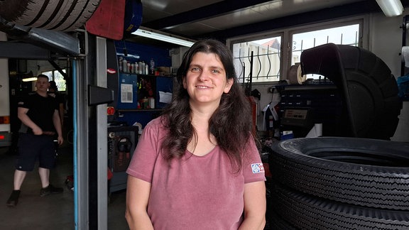 Eine Frau steht in einer Autowerkstatt und lächelt in die Kamera.
