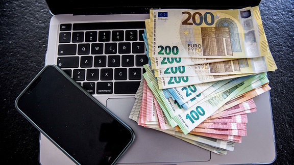 Mehrere tausend Euro in Bargeld liegen auf einen Laptop und daneben ein Handy. 