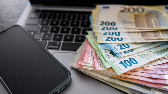 Mehrere tausend Euro in Bargeld liegen auf einen Laptop und daneben ein Handy. 