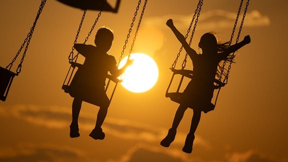 Kinder sitzen an der Strandpromenade in einem Kettenkarussell und zeichnen sich im Gegenlicht der untergehenden Sonne als Silhouette ab.