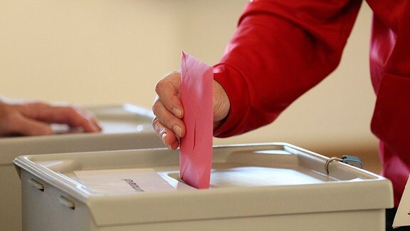 Mensch steckt Stimmzettel in eine Wahlurne.