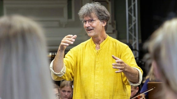 Ein Mann mit grauen Haaren, Brille, Schnurrbart und gelbem Hemd dirigiert ein Orchester.