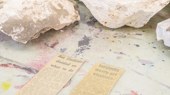 Steine liegen auf dem Boden neben alten ausgeschnittenen Zeitungsartikeln.