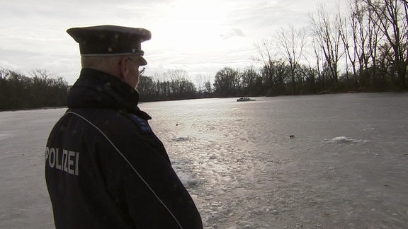 Ein Polizist mit Uniform und Jacke steht mit dem Rücken zur Kamera und blickt auf eine gefrorene Wiesenfläche. Ganz hinten sieht man ein Auto im Eis stecken.