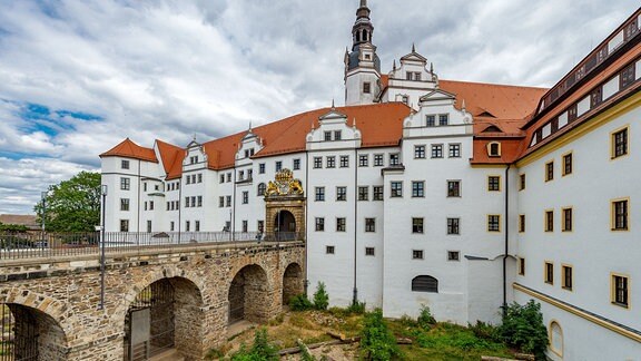 Schloss Hartenfels, im Vordergrund eine steinerne Brücke, die auf ein mehrstöckiges Gebäude mit weißer Fassade zuführt.