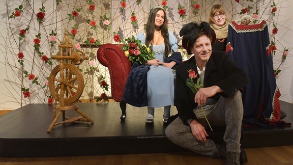 Zwei Frauen und ein Mann posieren für ein Foto auf einer Bühne mit einem roten Sofa und einem Spinnrad darauf, im Hintergrund hängen zahlreiche Rosen an der Wand.