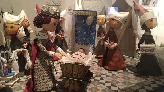 Eine Vitrine zeigt eine Reihe von Puppen in mittelalterlicher Kleidung, die um eine weitere Puppe mit Krone, Kleid und rotem Umhang neben einer Wiege stehen