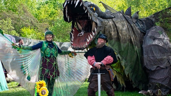 Zwei Menschen in einem Ritter und einem Elfenkostüm stehen vor einer Drachenskulptur.