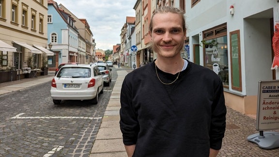 Ein junger Mann steht in einer Straße in der Grimmaer Innenstadt und lächelt in die Kamera. 