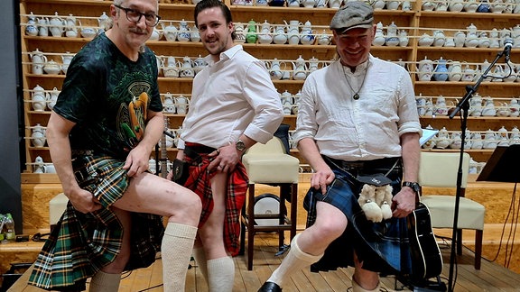 Drei Männer der Live-Band Leprechaun's Pleasure in schottischen und irischen Röcken stehen auf einer Bühne und ziehen den Rock jeweils etwas höher über ihre Knie. Sie lachen in die Kamera, verraten aber nicht, was sich unter diesen Röcken befundet. 