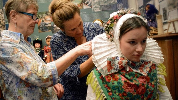 Zwei Frauen gestalten bei einer dritten Frau ihr sorbisches Kostüm.