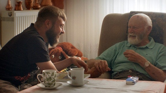 Zwei Männer an einem Kaffeetisch, der Großvater im Sessel, sein Enkel neigt sich ihm zu.
