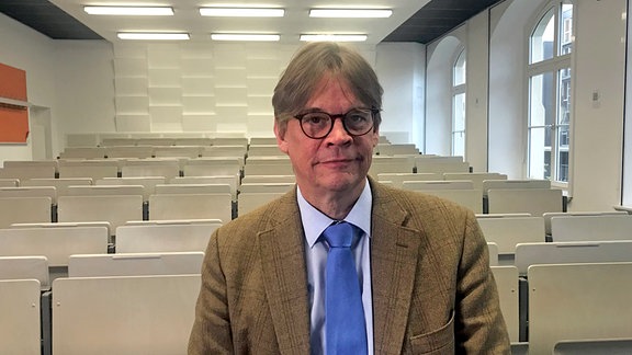 Professor Harald Simons, ein Mann mit Brille und braunen Haaren steht vor Stuhlreihen und schaut in die Kamera