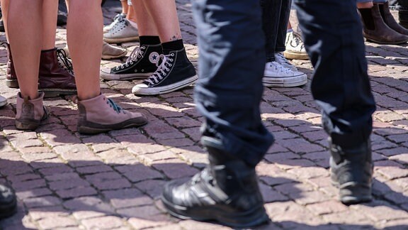 Symbolbild von Füßen in Turnschuhen auf Kopfsteinpflaster. Im Vordergrund: Schwarze Uniformhosen in Stiefeln.
