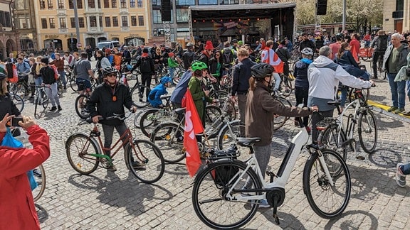 Dutzende Menschen mit Fahrrädern stehen auf einem Platz in der Sonne. Es sind die Teilnehmer einer Fahrraddemonstration zum 1. Mai 2023 in Leipzig auf dem Marktplatz.