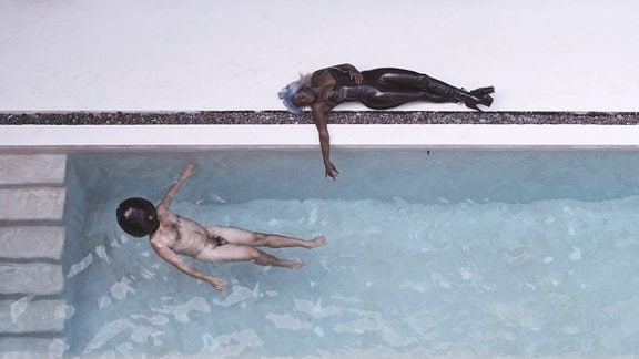 Ein Schwimmbassin in dem ein nackter Mann mit schwarzer Maske schwimmt, am Rand liegt eine schwarze Frau in Latexsachen.