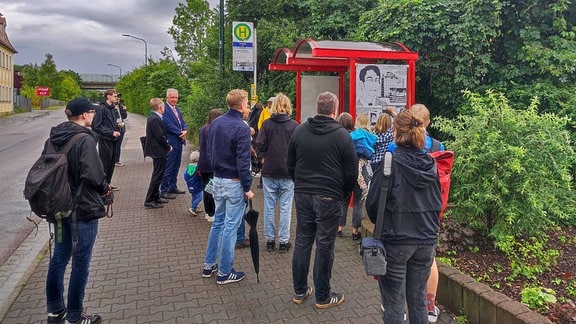 Mehr als ein Dutzend Menschen stehen vor einer Bushaltestelle, an deren Wänden Zeichnungen eines jungen Mannes geklebt sind