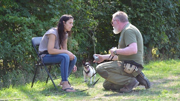 Ein Mann und eine Frau sitzen auf einer Wiese, daneben steht ein kleiner Hund.