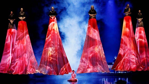 Mehrere Figuren, die meterhoch über der Bühne schweben, unter sich lange Stoffbahnen wie Röcke.