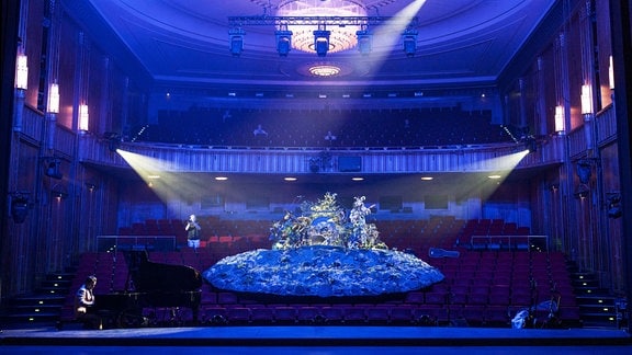 Zuschauerraum eines Theaters, in der Mitte ist eine große Insel auf den Stuhlreihen zu sehen, darin ein Schlagzeuger, an anderen Stellen des Zuschauerraumes sind weitere Musiker.