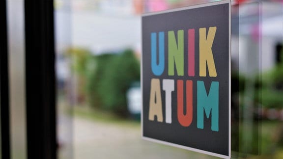 An einem Fenster ist ein Schild, auf dem in bunten Großbuchstaben das Wort "Unikatum" steht. 