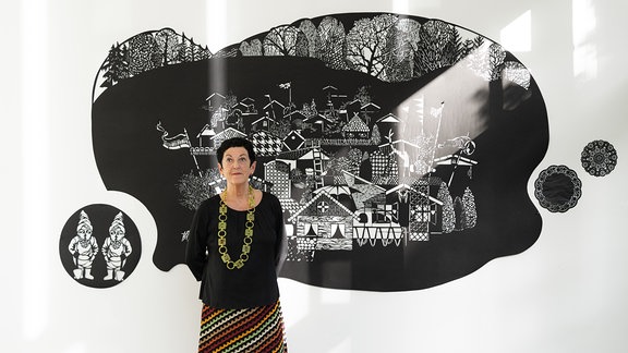 Eine Frau mit kurzen dunklen Haaren, dunklem Pullover und bunt gestreiftem Rock steht vor einem schwarz-weißen Kunstwerk an einer Wand. Darauf sind kleine Häuser und Gartenzwerge zu sehen.