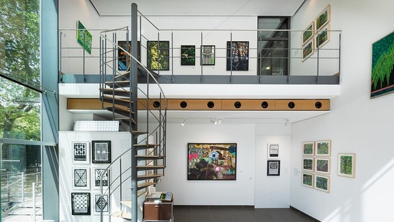 Blick in einen hellen Ausstellungsraum mit Wendeltreppe. An den Wänden hängen mehrere Kunstwerke in bunten Farbtönen oder Schwarz-Weiß-Optik.