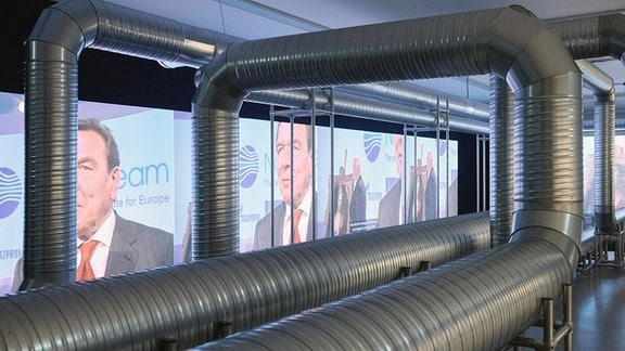 Große, metallische Rohre winden sich in einem Ausstellungraum vor einer Videoleinwand.